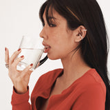 a sideview of lady with highlight hair, drinking a clear glass of water with red nailed hand, with w0dd ear cuffs and a red cardigan on