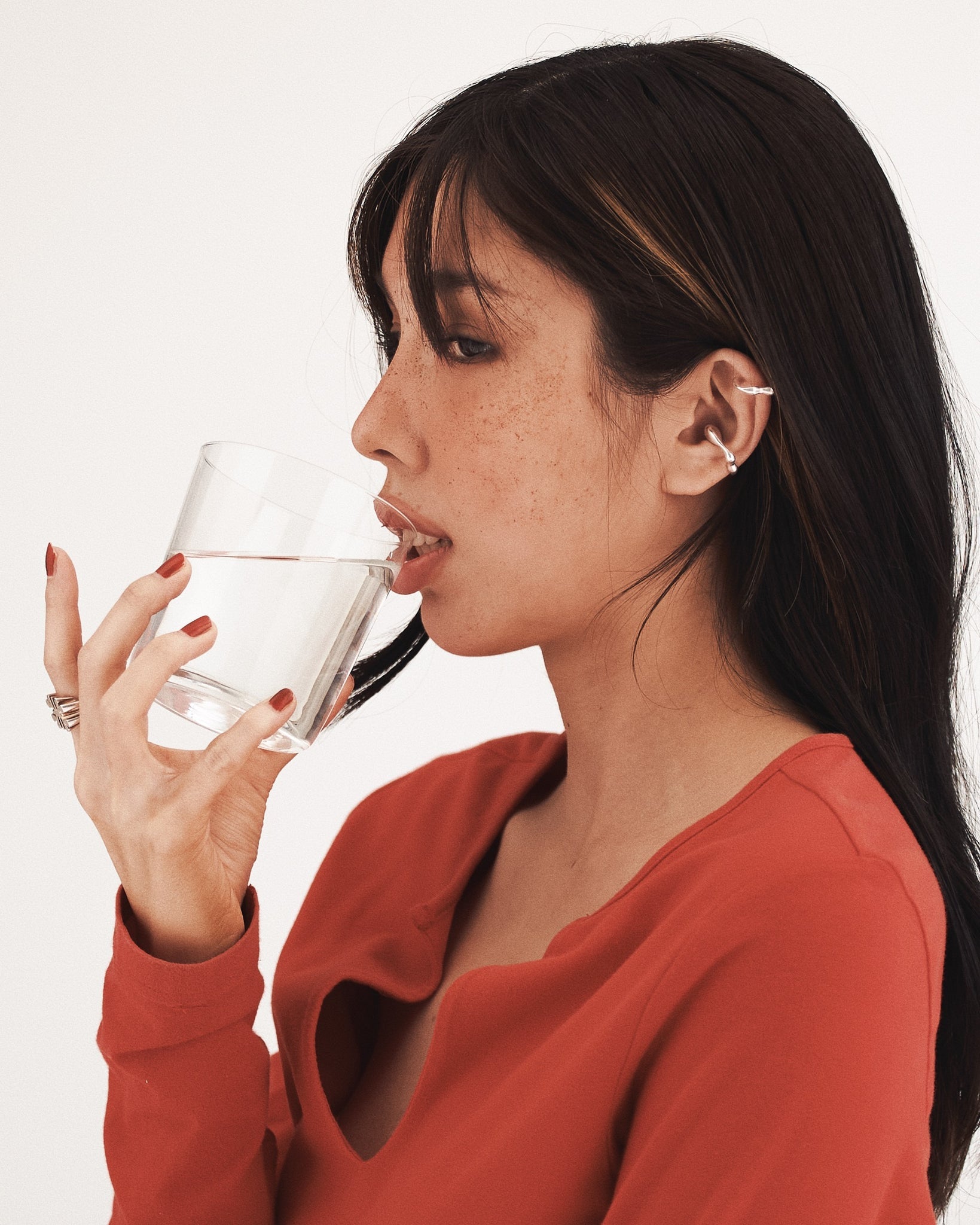 a sideview of lady with highlight hair, drinking a clear glass of water with red nailed hand, with w0dd ear cuffs and a red cardigan on