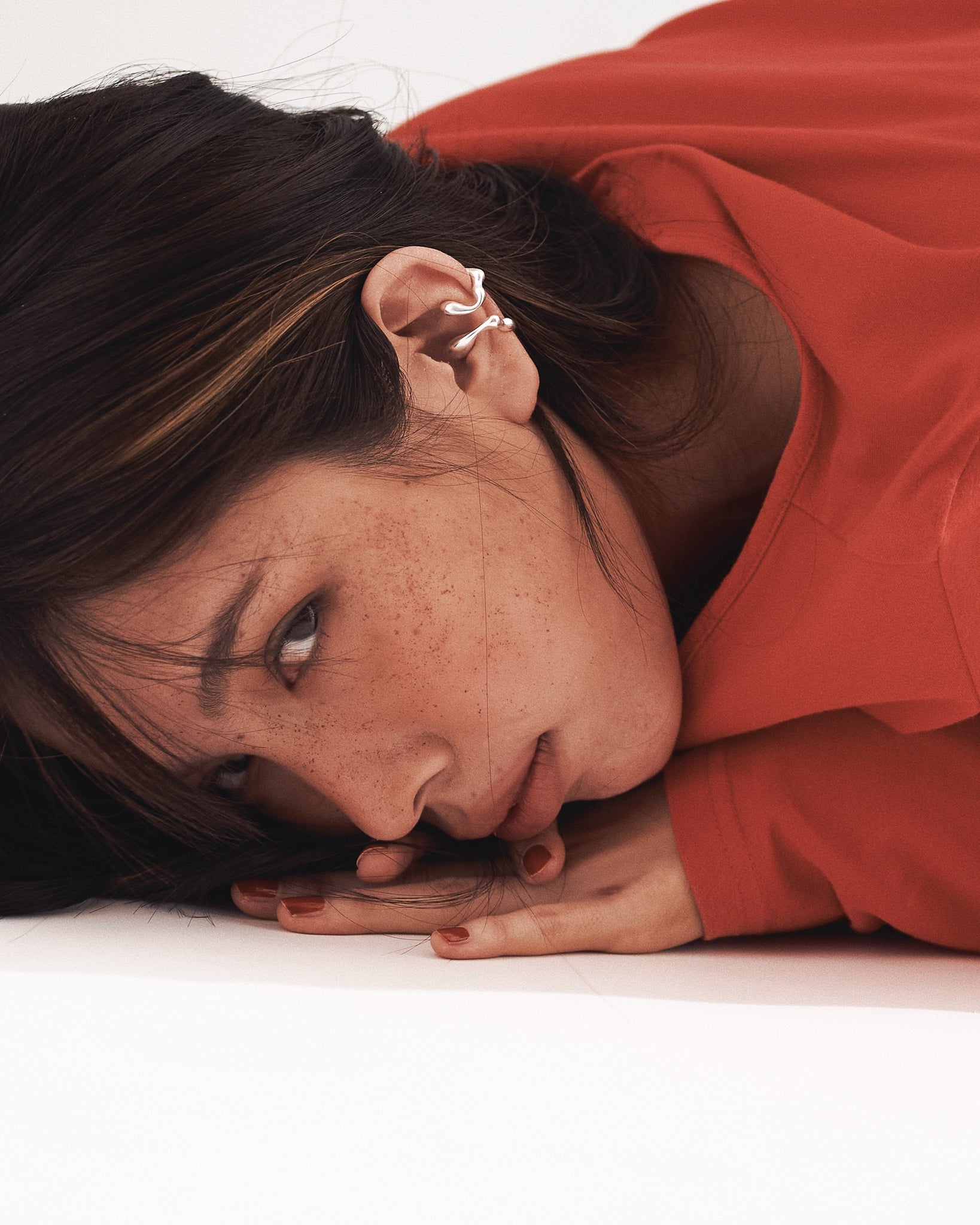 a lady with highlight hair, wearing 2 w0dd ear cuffs on one ear and laying face down on a white floor